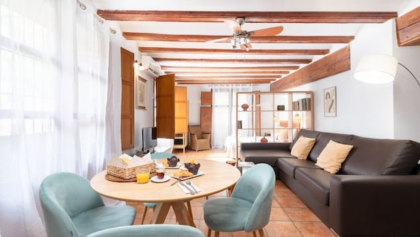 A cozy living room with a round wooden table, two light blue chairs, a dark brown sofa, and a ceiling fan.