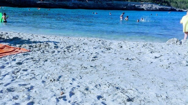 Ein Strand mit klarem, blauem Wasser, sandigem Ufer und felsigen Klippen im Hintergrund.