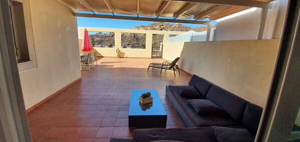 A covered patio with a sofa, a table, and chairs. There is a view of a mountainous landscape in the background.