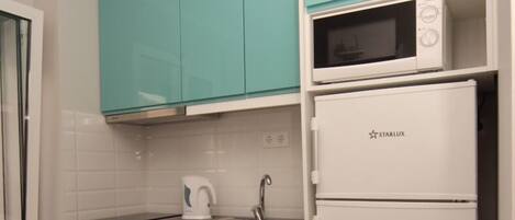 Een kleine keuken met blauwgroene kastjes, een wasmachine, een magnetron en een koelkast.