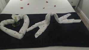 En säng med en svart- och vitrandig kudde, en röd kudde och en hjärtformad handduk som är vackert arrangerad på ett svart sängöverkast.