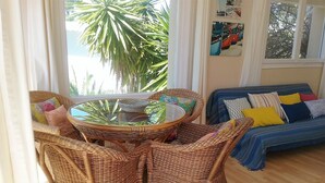 Ein Zimmer mit einem Korbspeiseset, einer Couch mit bunten Kissen und Blick auf Palmen durch das Fenster.