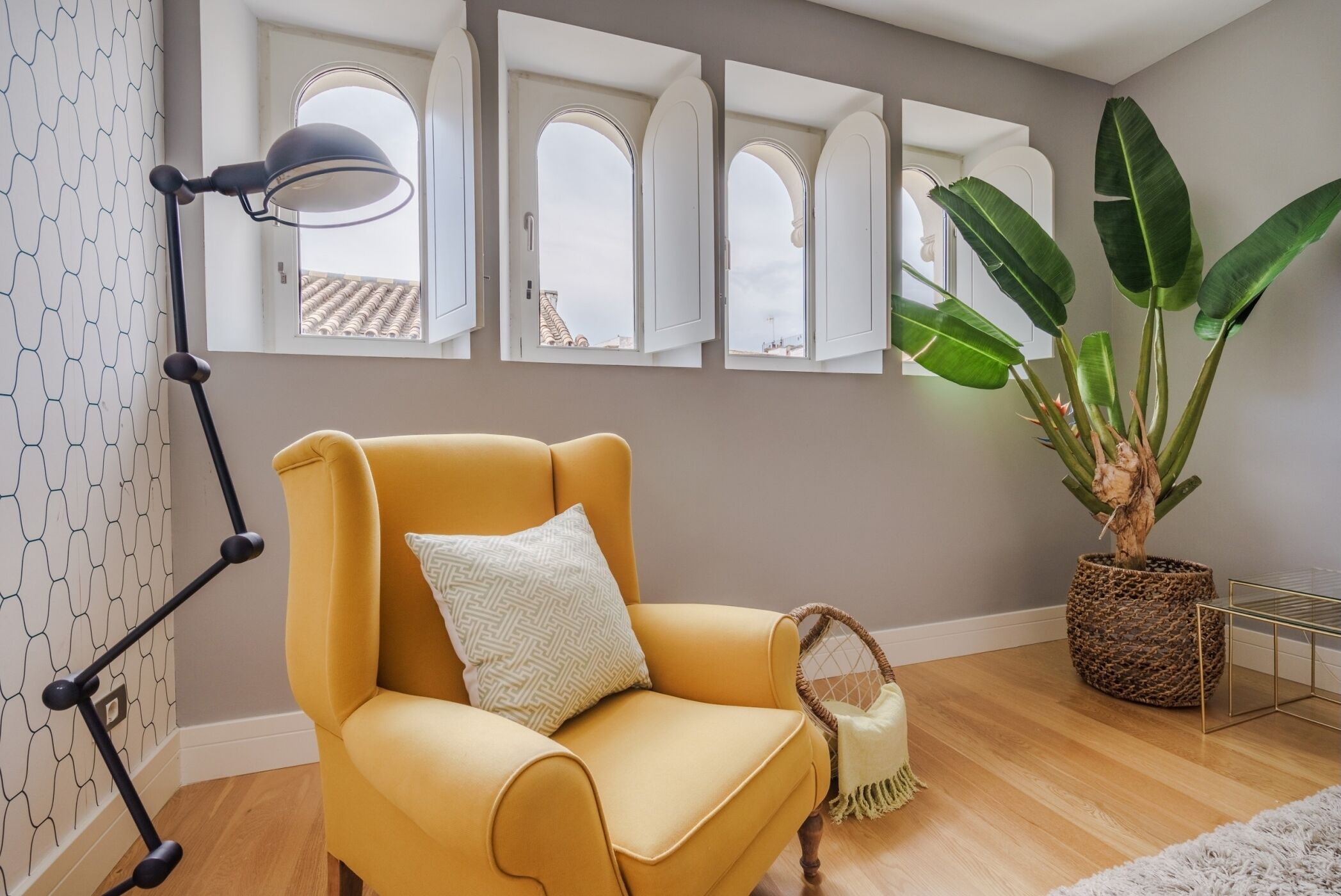 A room with a yellow armchair, a large potted plant, a floor lamp, and a view through arched windows.