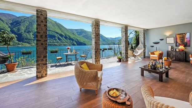A spacious indoor area with a view of a lake and mountains, furnished with wicker chairs and a dining table.
