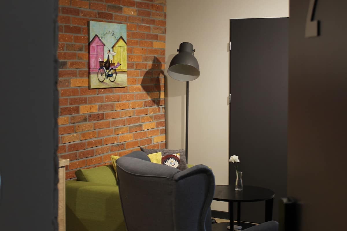 Un salon moderne avec un mur en briques, un canapé vert, un fauteuil foncé, une petite table ronde et un lampadaire.