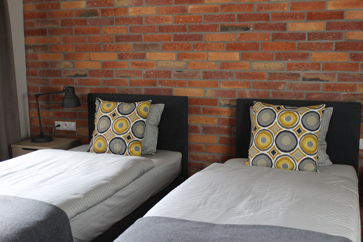 Une chambre avec deux lits, chacun agrémenté d’un coussin à motifs, le tout adossé à un mur de briques.
