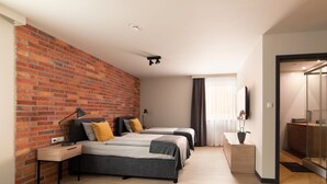A modern hotel room with a brick accent wall, two beds, a desk, a TV, and a bathroom with a glass shower.