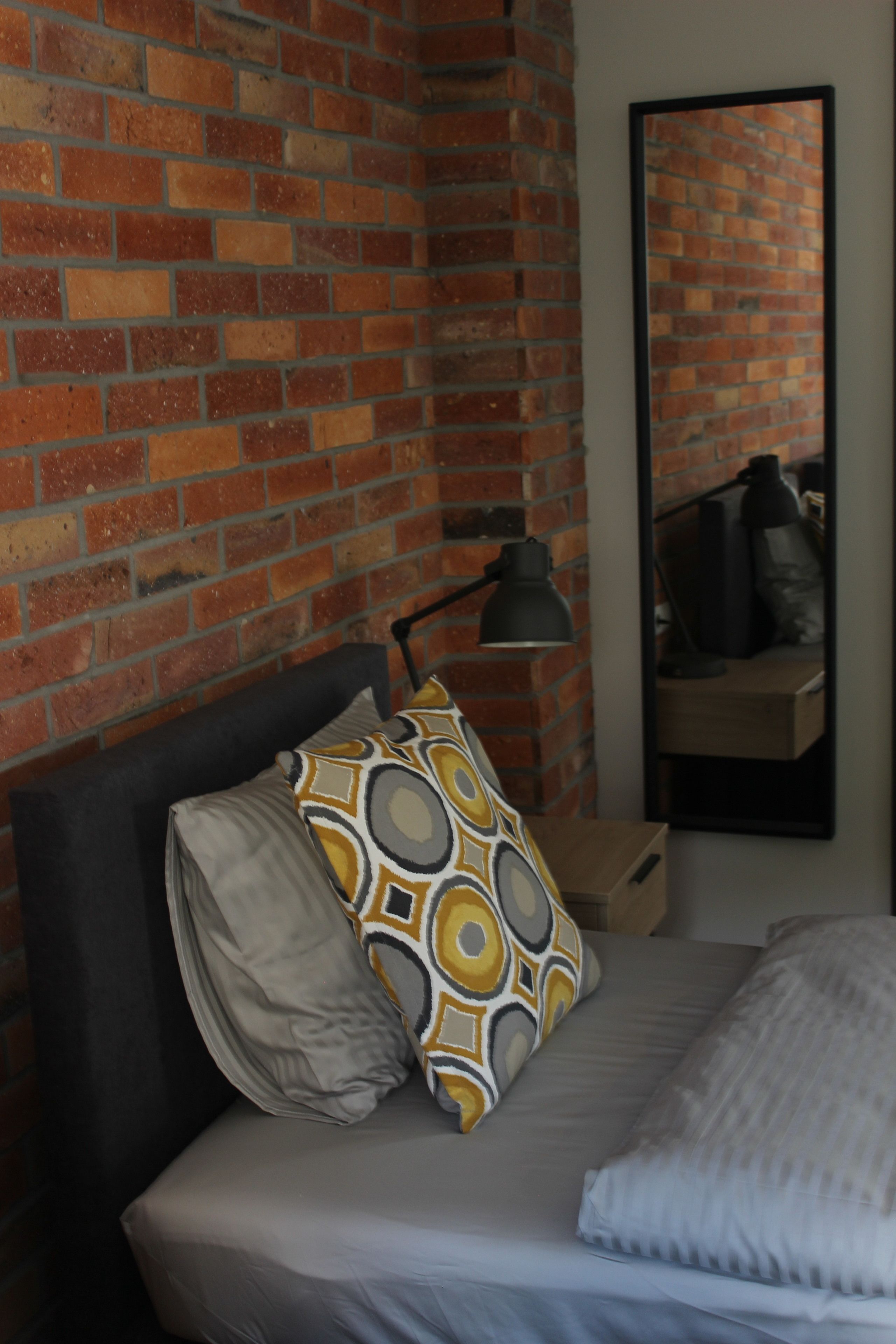 Un dormitorio con pared de ladrillo, cama con almohadas estampadas, mesita de noche con lámpara y espejo en la pared.