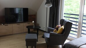 A modern living room with a flat-screen TV, a round table with a vase, a grey armchair with a patterned cushion, and a large window with blinds.