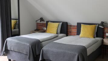 A bedroom with two beds, each with a grey bedspread and a yellow pillow. There is a nightstand with a lamp on each side of the beds. The room has a brick wall and a large mirror on the left.