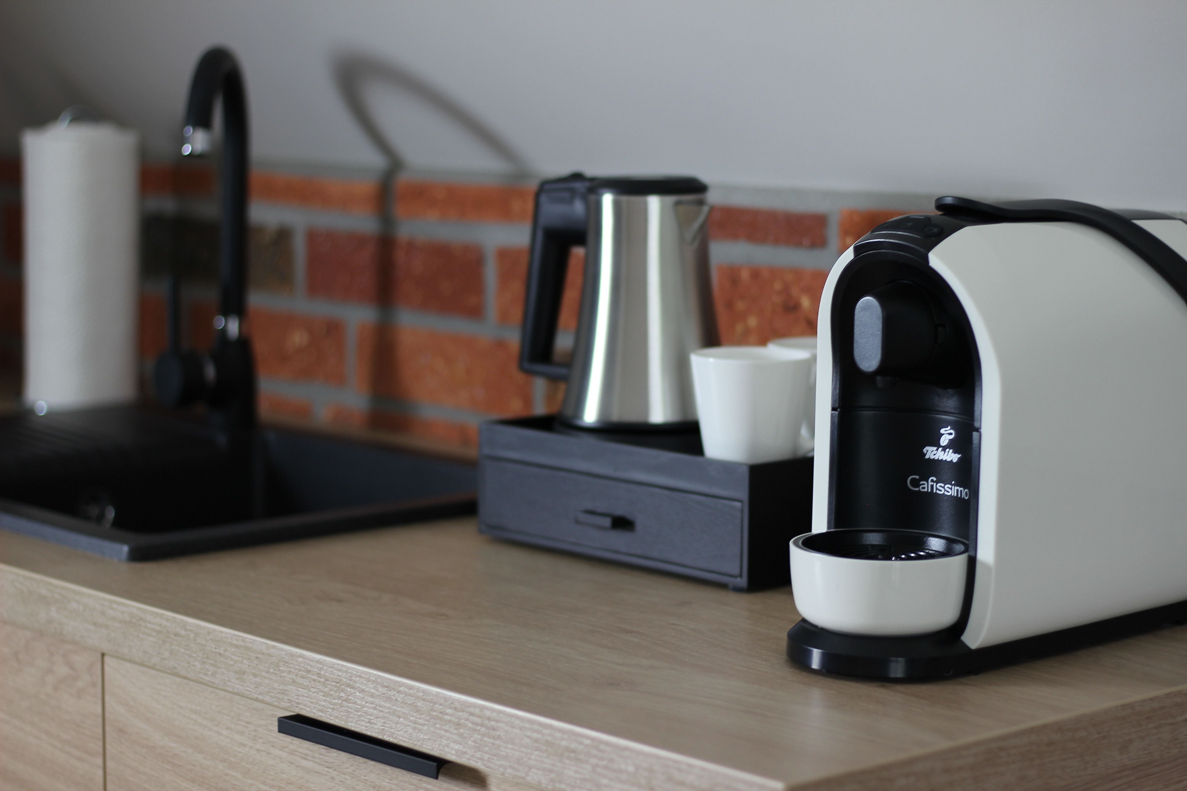 Eine Küche mit Kaffeemaschine, Wasserkocher und Tassenspender auf einer Holzausziehtischplatte.