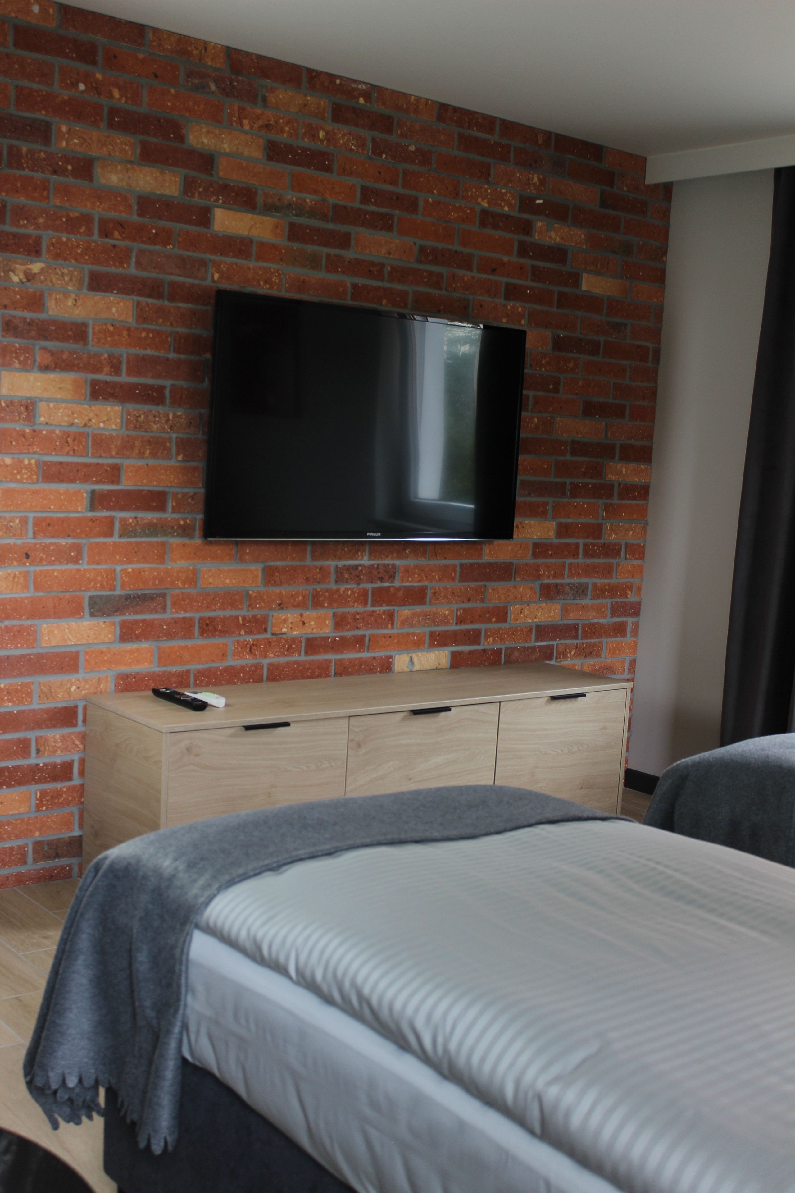 A hotel room with a brick wall, a flat-screen TV mounted above a wooden dresser, and two beds with grey bedding.