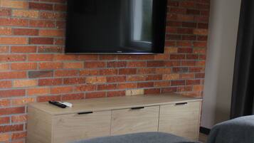 A hotel room with a brick wall, a flat-screen TV mounted above a wooden dresser, and two beds with grey bedding.