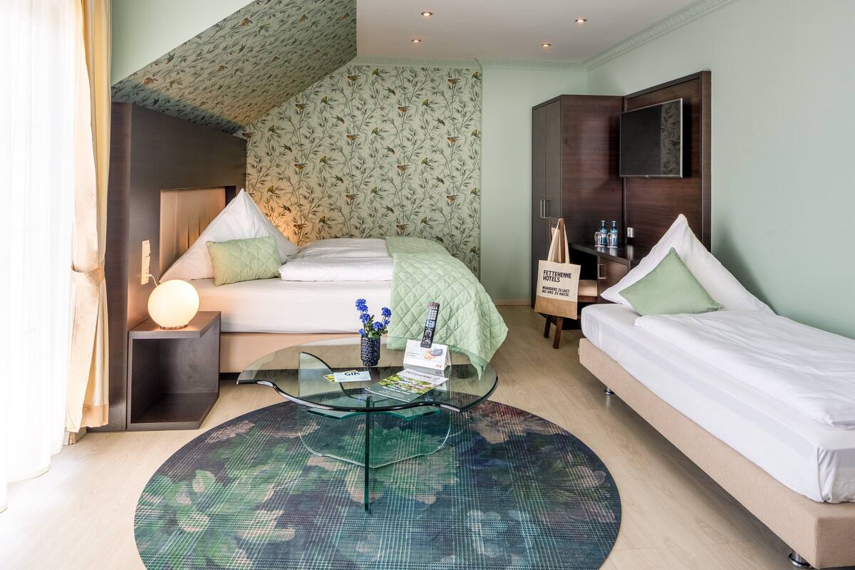 A hotel room with a bed, a bedside table with a lamp, a glass coffee table, a small table with a vase of flowers, and a wall-mounted TV.