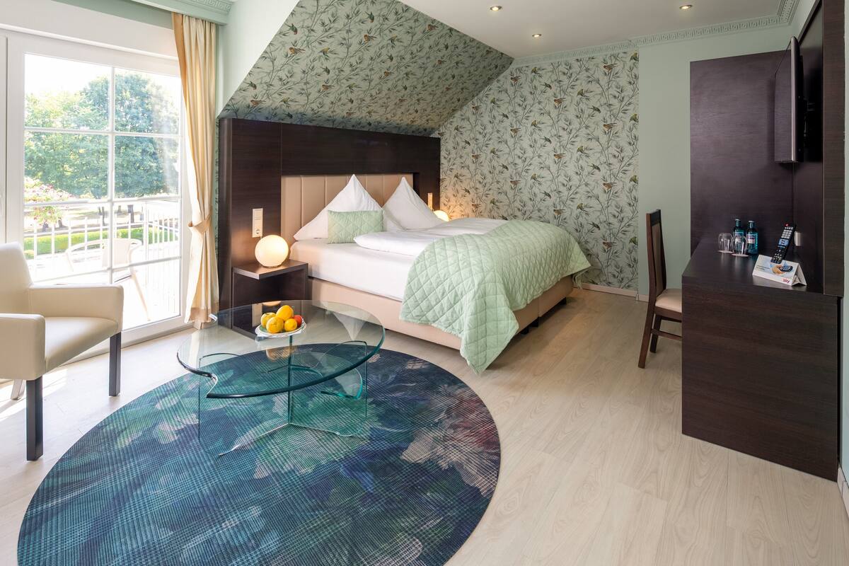 A modern hotel room with a large bed, a glass coffee table with fruit, a seating area with a chair, and a view of the outdoors.