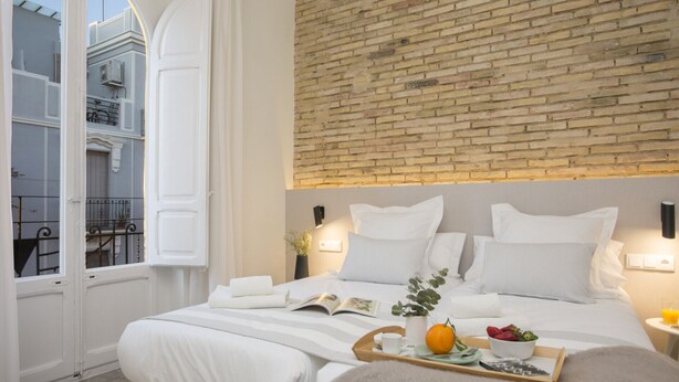 Une chambre d’hôtel avec un mur d’accent en briques, un grand lit avec une literie blanche, une table de chevet sur laquelle se trouve un plateau de fruits et un livre, et une vue sur un balcon où se trouve un vélo.