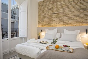 Une chambre d’hôtel avec un mur d’accent en briques, un grand lit avec une literie blanche, une table de chevet sur laquelle se trouve un plateau de fruits et un livre, et une vue sur un balcon où se trouve un vélo.