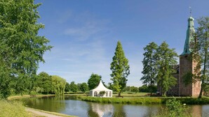A historic building with a tall tower and a small pavilion by a pond. - Hotel am Tiergarten (Raesfeld)