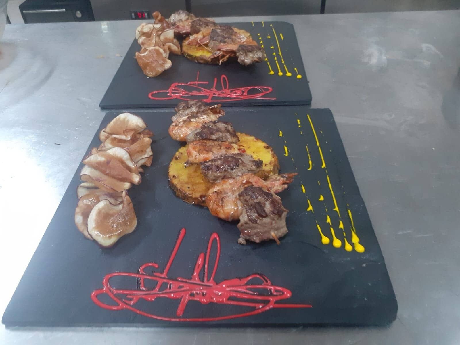 Een eettafel met een zwarte arduinen blad, met diverse soorten eten, waaronder gegrild vlees, paddenstoelen en aardappelen, en rode en gele decoratieve elementen.