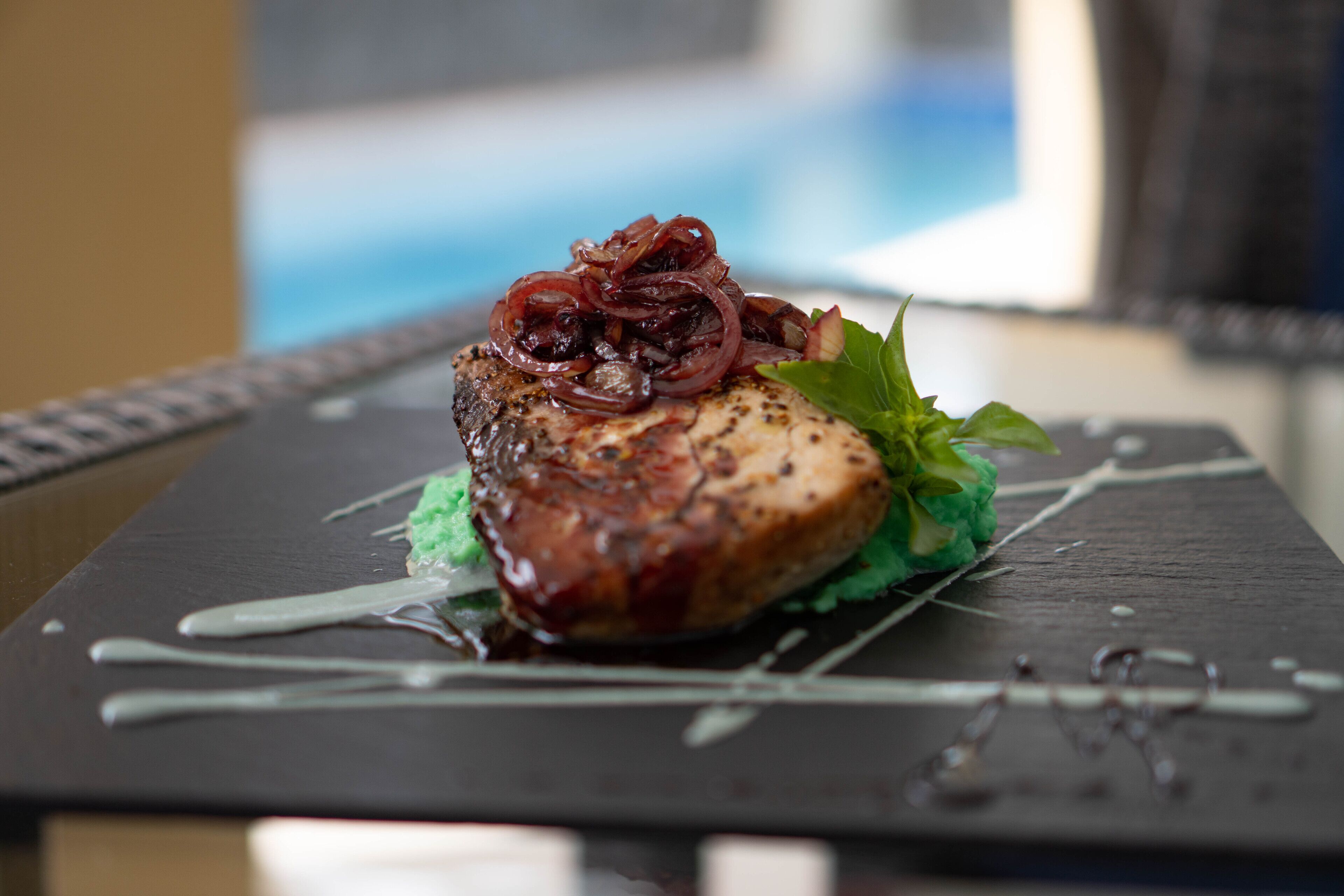 A plated dish with a piece of meat, garnished with red onions and a green leaf, on a dark square plate.