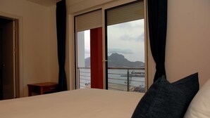 Ein Hotelzimmer mit Bett, einem kleinen Tisch, einer roten Akzentwand und Blick auf Berg und Wasser durch ein großes Fenster.