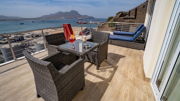 Ein Balkon mit Korbmöbeln, einem Glastisch und Blick auf das Meer und die Berge.