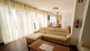 Ein Wohnzimmer mit einem beigefarbenen Sofa, einem Couchtisch aus Holz mit einem Zettel und Blick auf das Meer durch große Fenster.