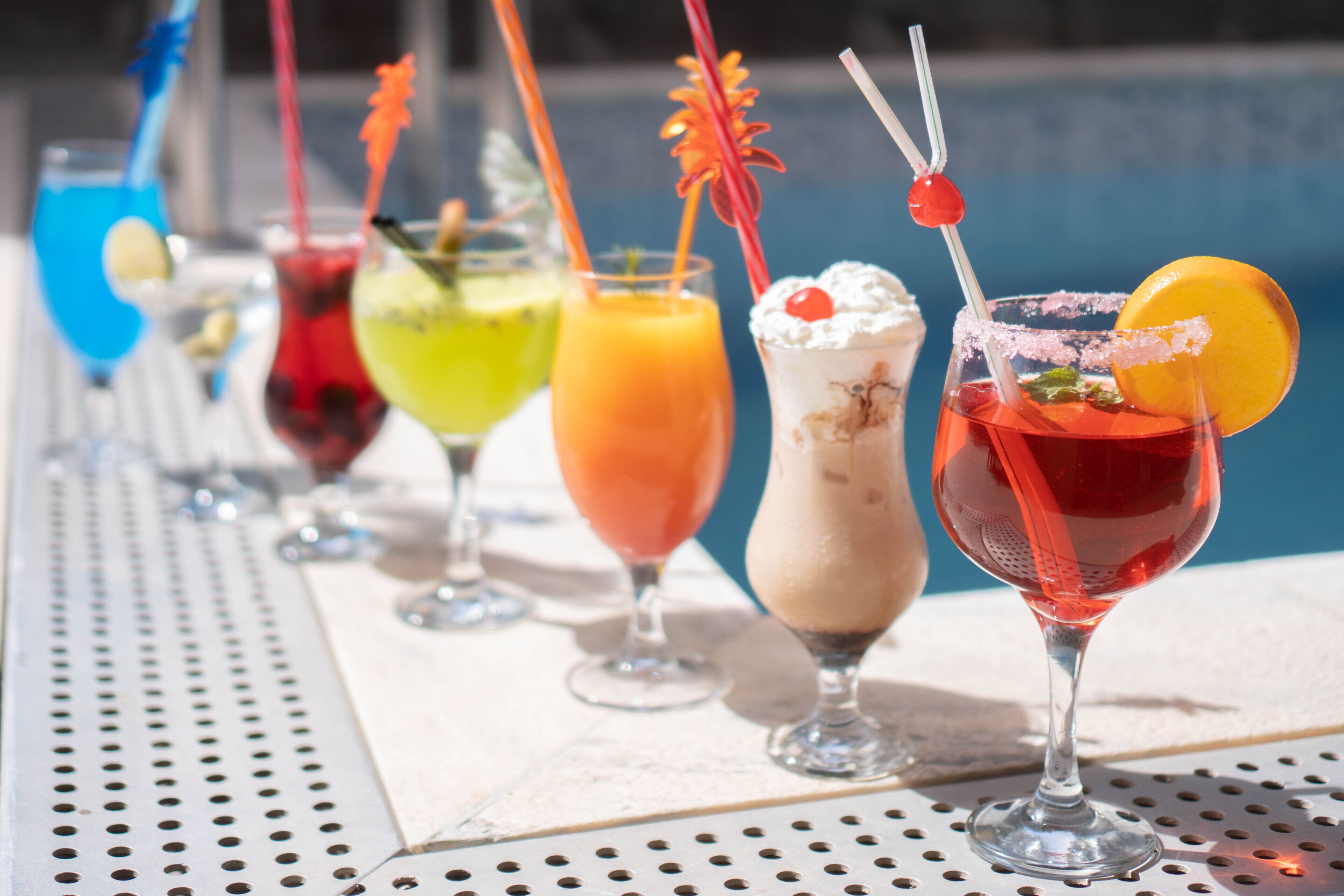 A row of colorful cocktails with various garnishes, placed on a table near a pool.