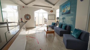 Eine moderne Hotelhalle mit Glastrennwand, einer Sitzecke mit dunklen Sofas und türkisfarbenen Kissen, einem Couchtisch mit Pflanze und einem großen Gemälde eines Segelbootes an der Wand.