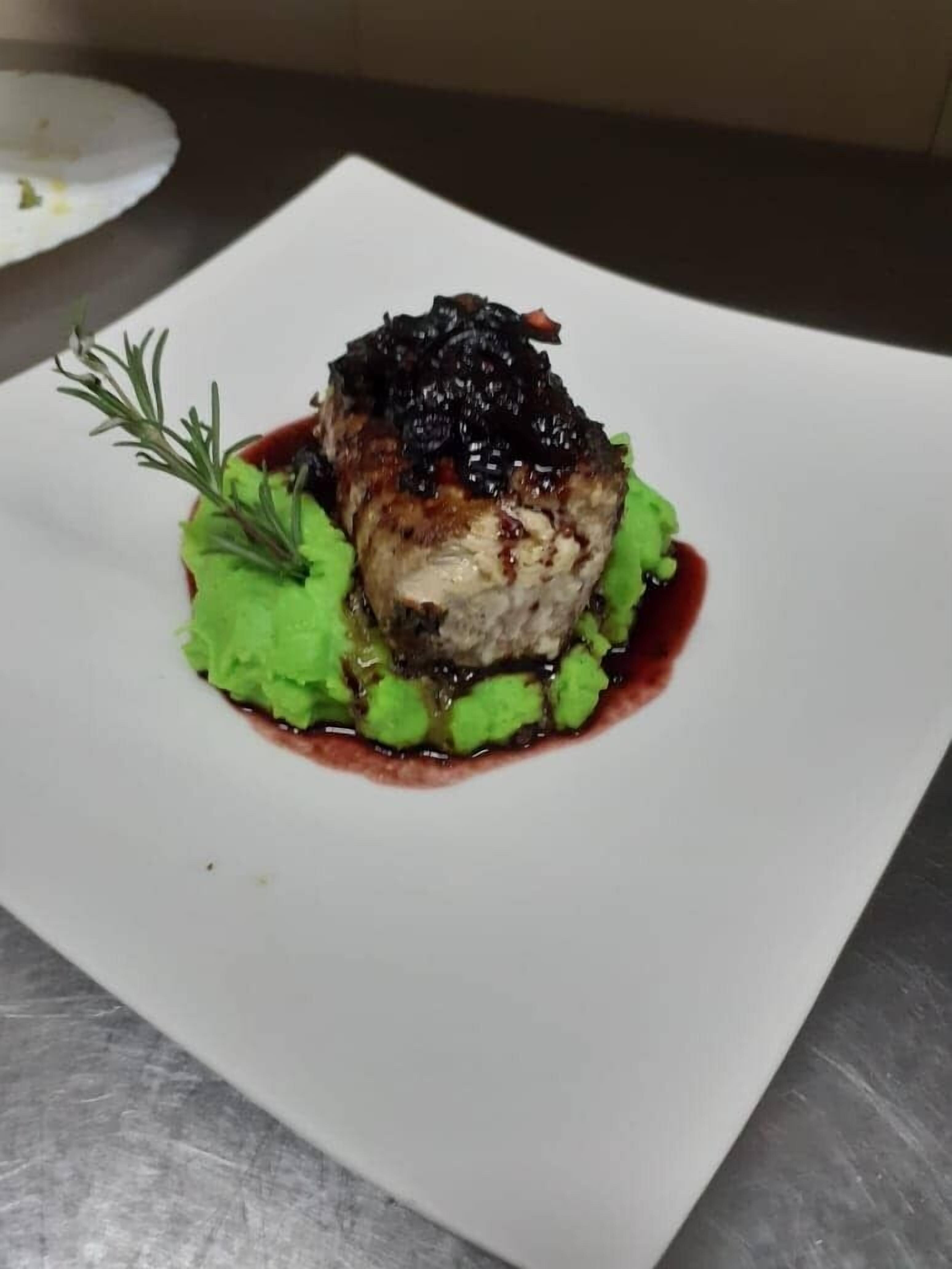A square plate with a piece of meat topped with a dark sauce, green puree, and a sprig of rosemary.