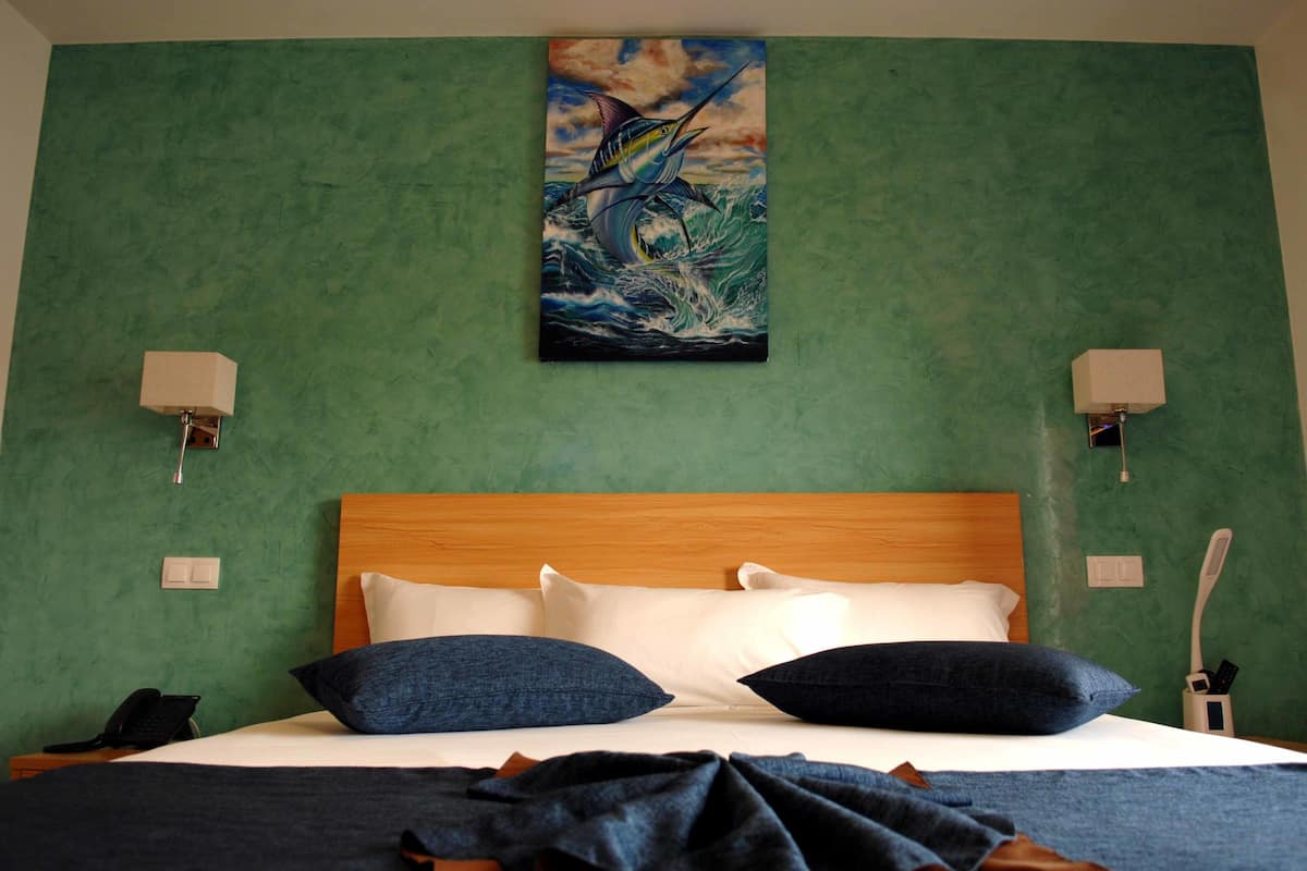 Une chambre d’hôtel avec un mur vert, une tête de lit en bois, deux oreillers blancs, deux oreillers bleu foncé, une couverture, un tableau représentant un poisson et deux lampes fixées au mur.