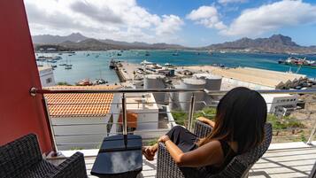 A balcony with wicker furniture overlooking a marina with boats and mountains in the background.