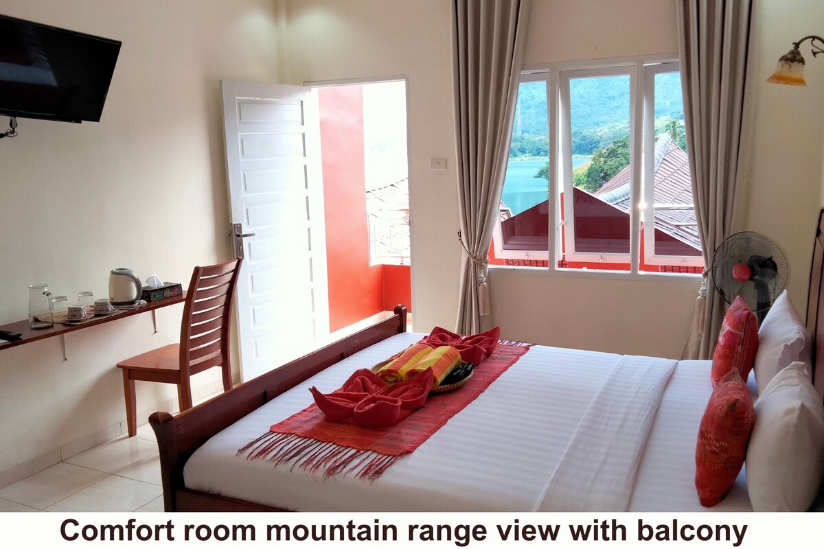 A bedroom with a mountain range view, a balcony, a bed with red bedding, a chair, a TV, and a fan.
