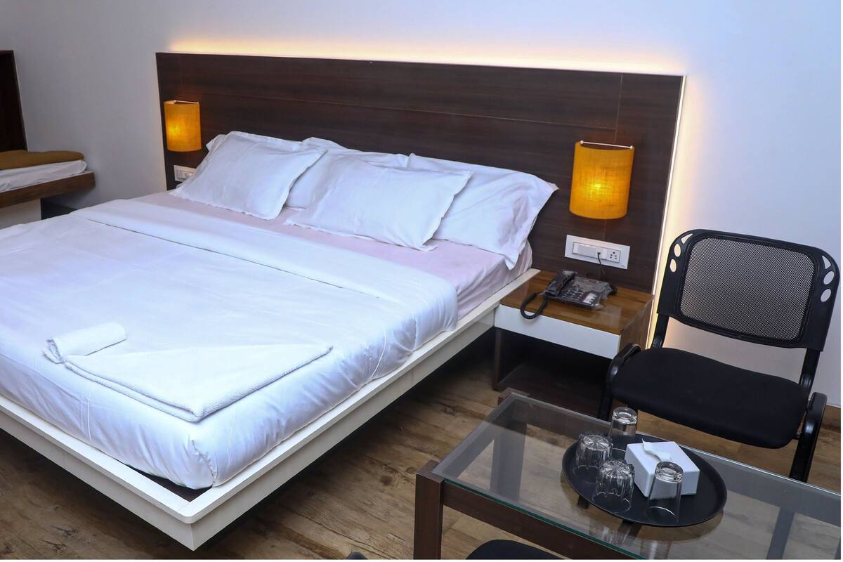 A neatly made bed with white linens, a bedside table with a lamp, a chair, and a glass table with a phone and a box.