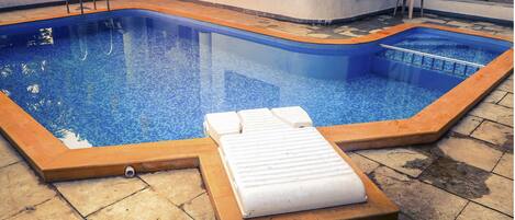 Una piscina rectangular con borde de azulejos y plataforma de madera. Hay sillas reclinables blancas y una toalla en la plataforma.