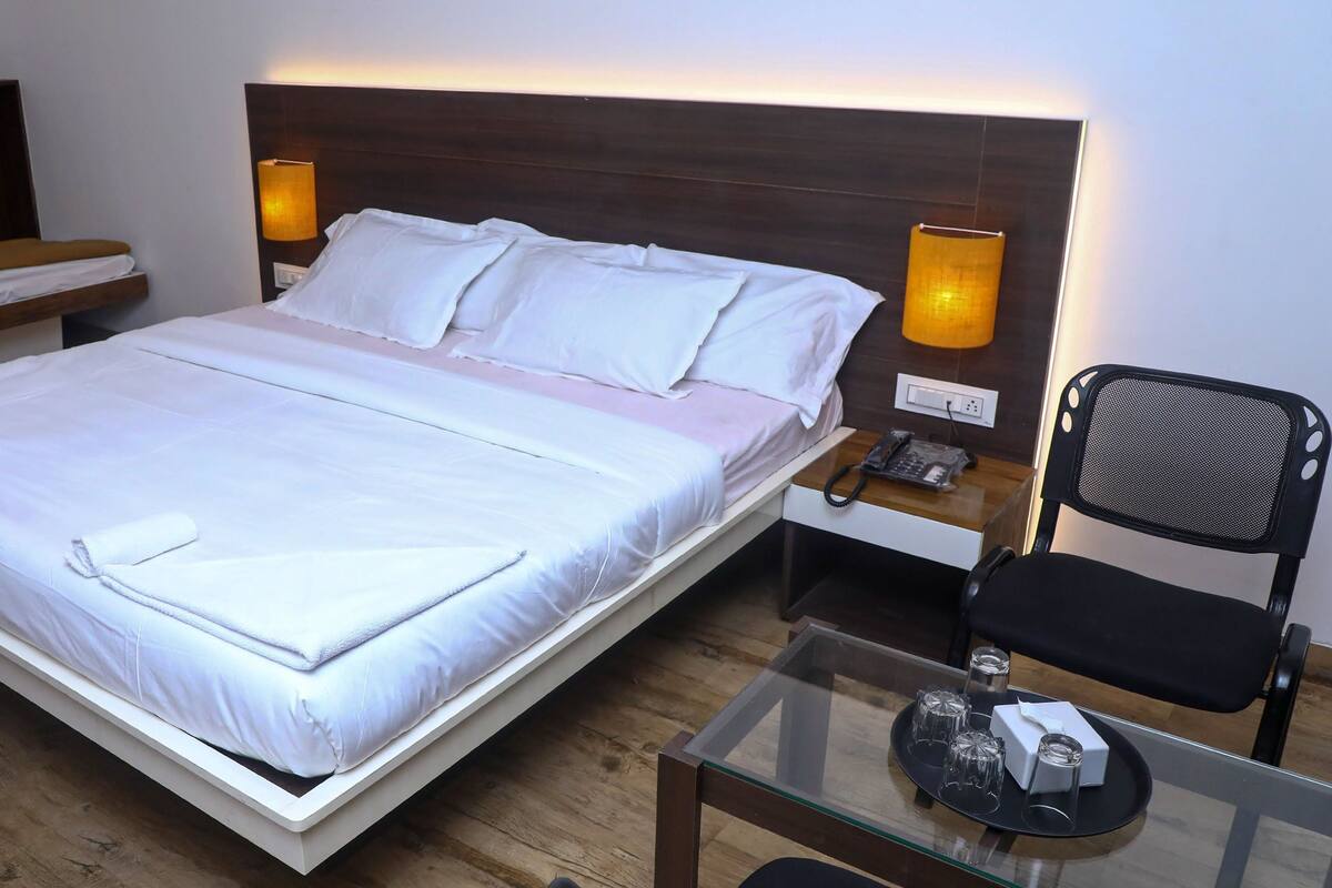 A hotel room with a large bed, bedside lamps, a desk with a phone, a chair, and a small table with glasses and a tissue box.