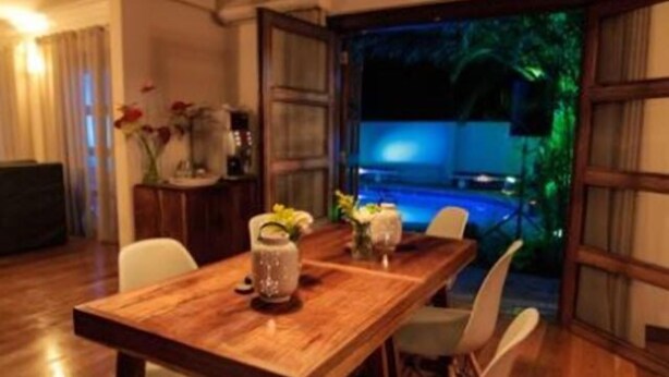 A dining area with a wooden table and white chairs, a hanging light fixture, and a view of a swimming pool at night. - MyGuest (Maputo)