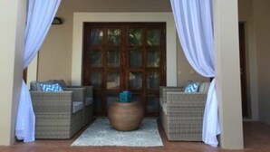 A room with a large wooden door, two wicker sofas with cushions, a round wooden table, and a white patterned rug. - MyGuest (Maputo)