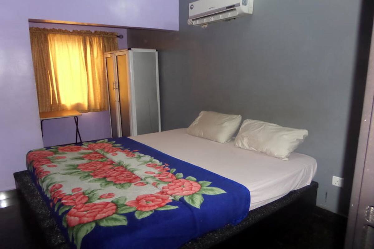 Un dormitorio con cama, mesita de noche, lámpara, ventana con cortinas y un equipo de aire acondicionado.