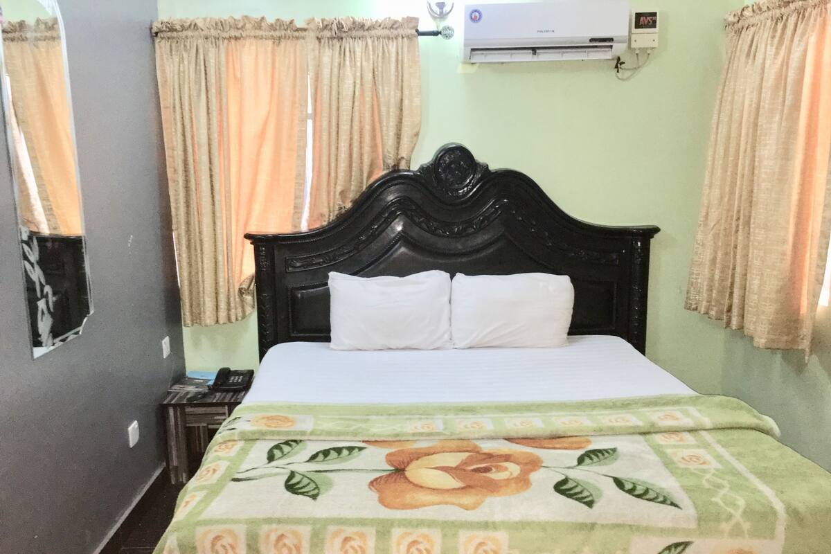 Una cama con colcha floral, cabecero negro y cortinas color beige.