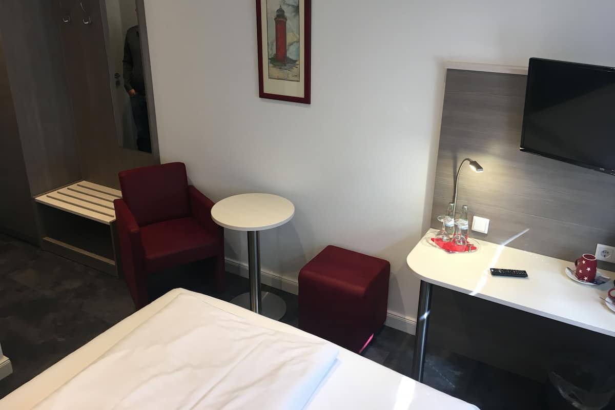 A hotel room with a bed, a red armchair, a small round table, a TV mounted on the wall, and a framed picture above the table.