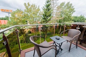 A balcony with two wicker chairs and a table, overlooking a garden with trees and a building in the background. - Hotel Am Stadtpark (Papenburg)
