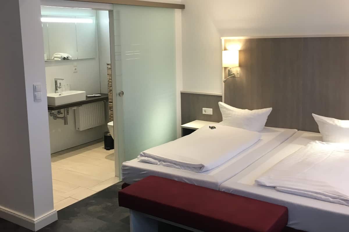 A hotel room with a bed, a bench, a nightstand, a mirror, a sink, and a toilet.