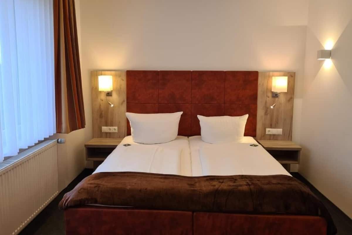 A double bed with a brown headboard, two bedside tables with lamps, and a window with curtains.