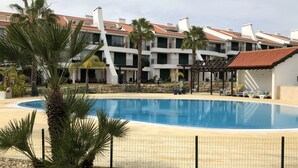 A swimming pool with a surrounding fence, palm trees, and a building with a tiled roof. - Luxury, spacious, two bedroom apartment on the Vill Sol Golf & Spa Resort (Quarteria)
