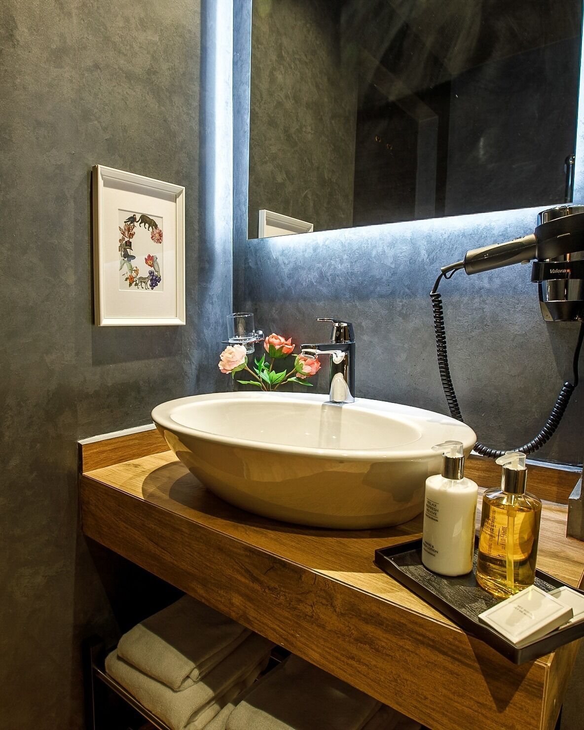 Un baño moderno con un lavamanos ovalado grande, un mueble de tocador de madera, una obra de arte enmarcada y un secador de pelo en la encimera.