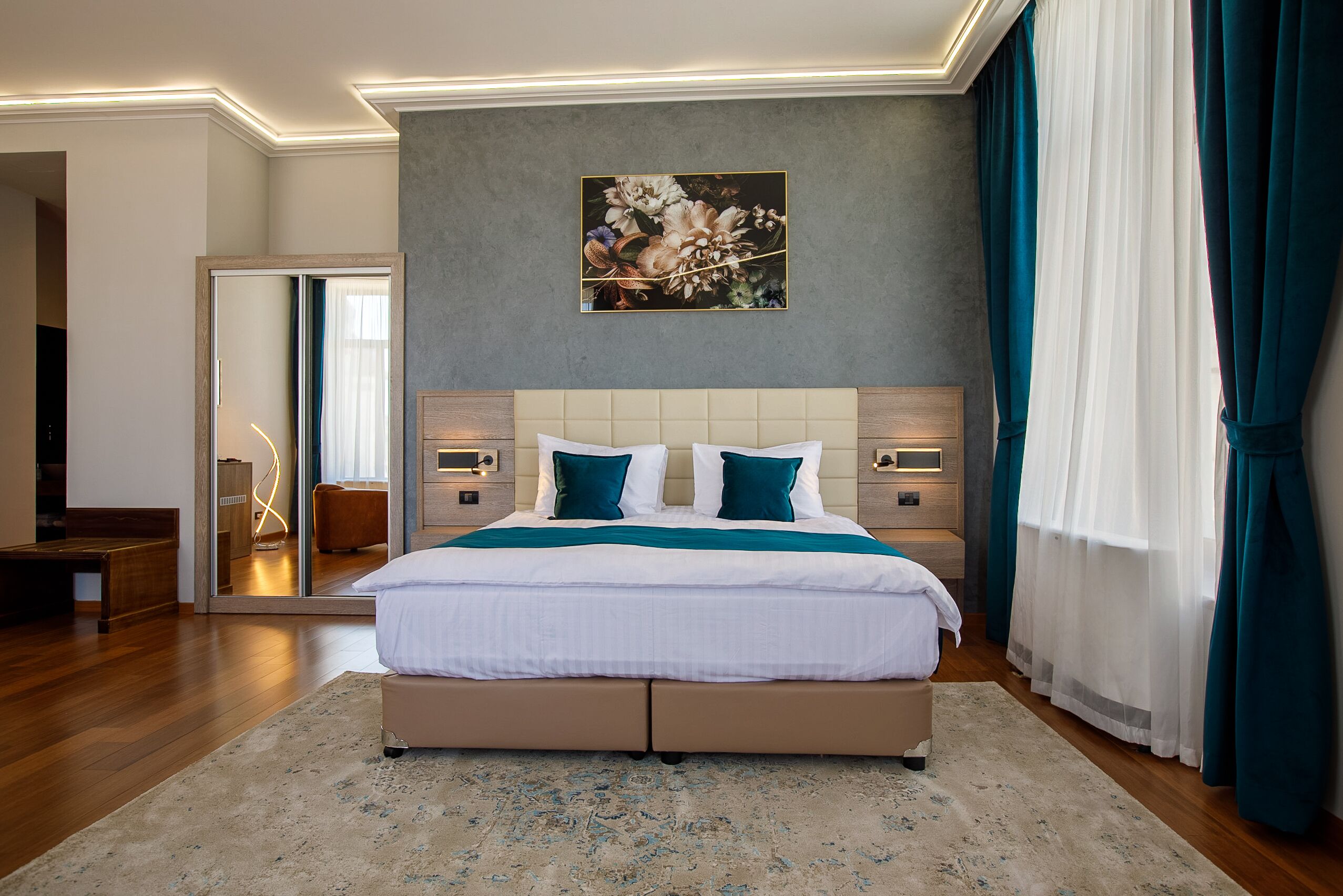 A modern bedroom with a large bed, a nightstand, a bedside table, a wall-mounted lamp, and a painting.