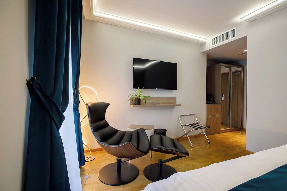 Une chambre d’hôtel moderne avec un fauteuil noir aux lignes courbes, un téléviseur à écran plat, une petite plante et un meuble en bois.