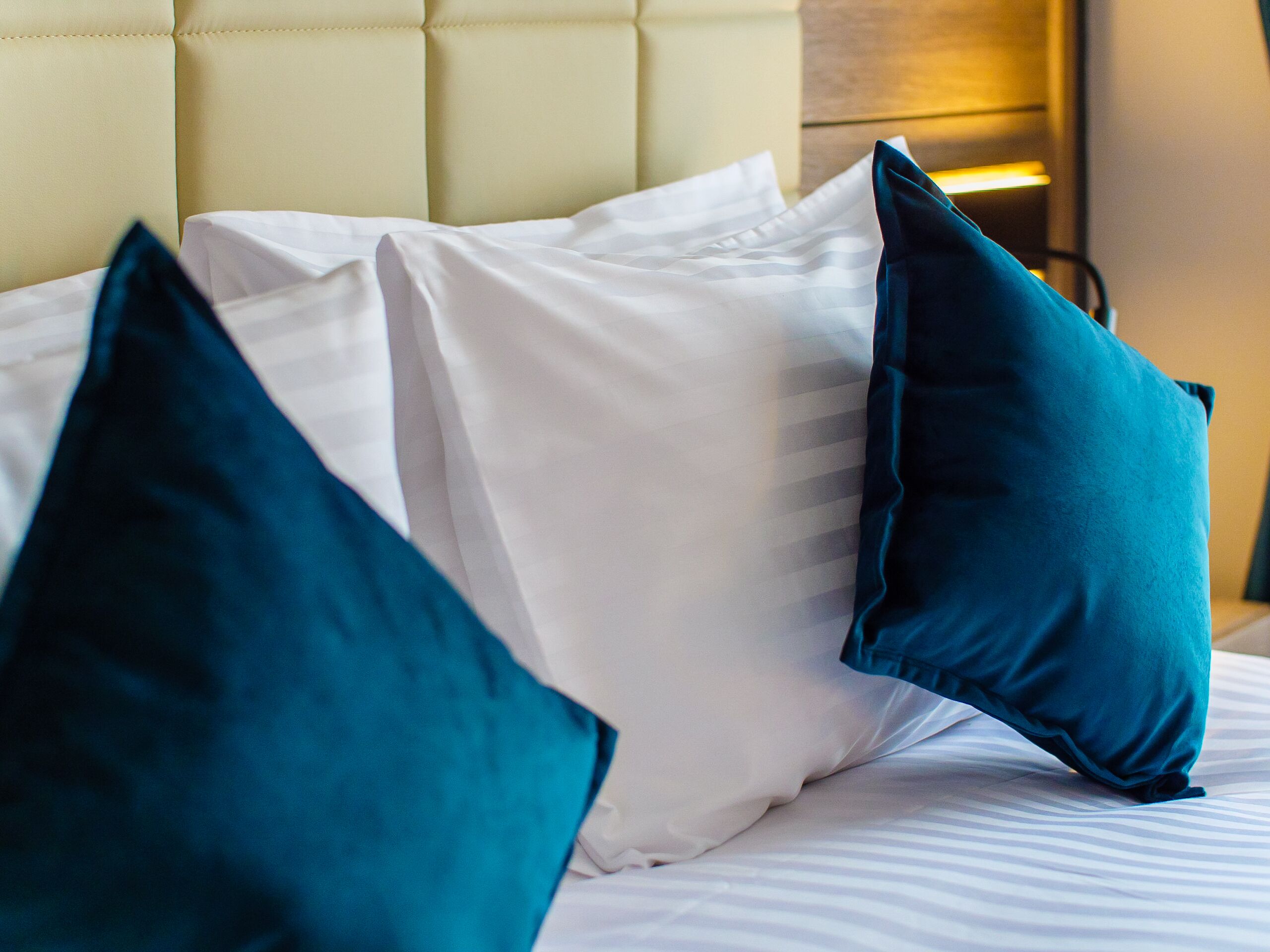 Un lit avec des oreillers blancs et turquoise, une tête de lit capitonnée et une table de chevet avec une lampe.