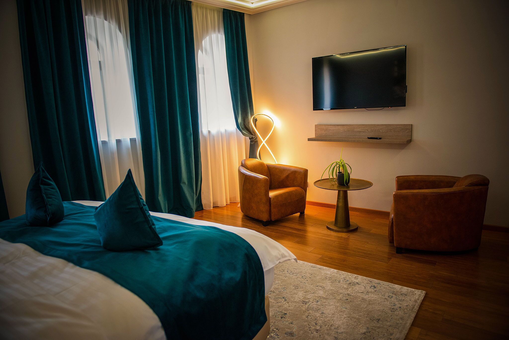 A hotel room with a bed, a TV mounted on the wall, a round table with a plant, and two armchairs.